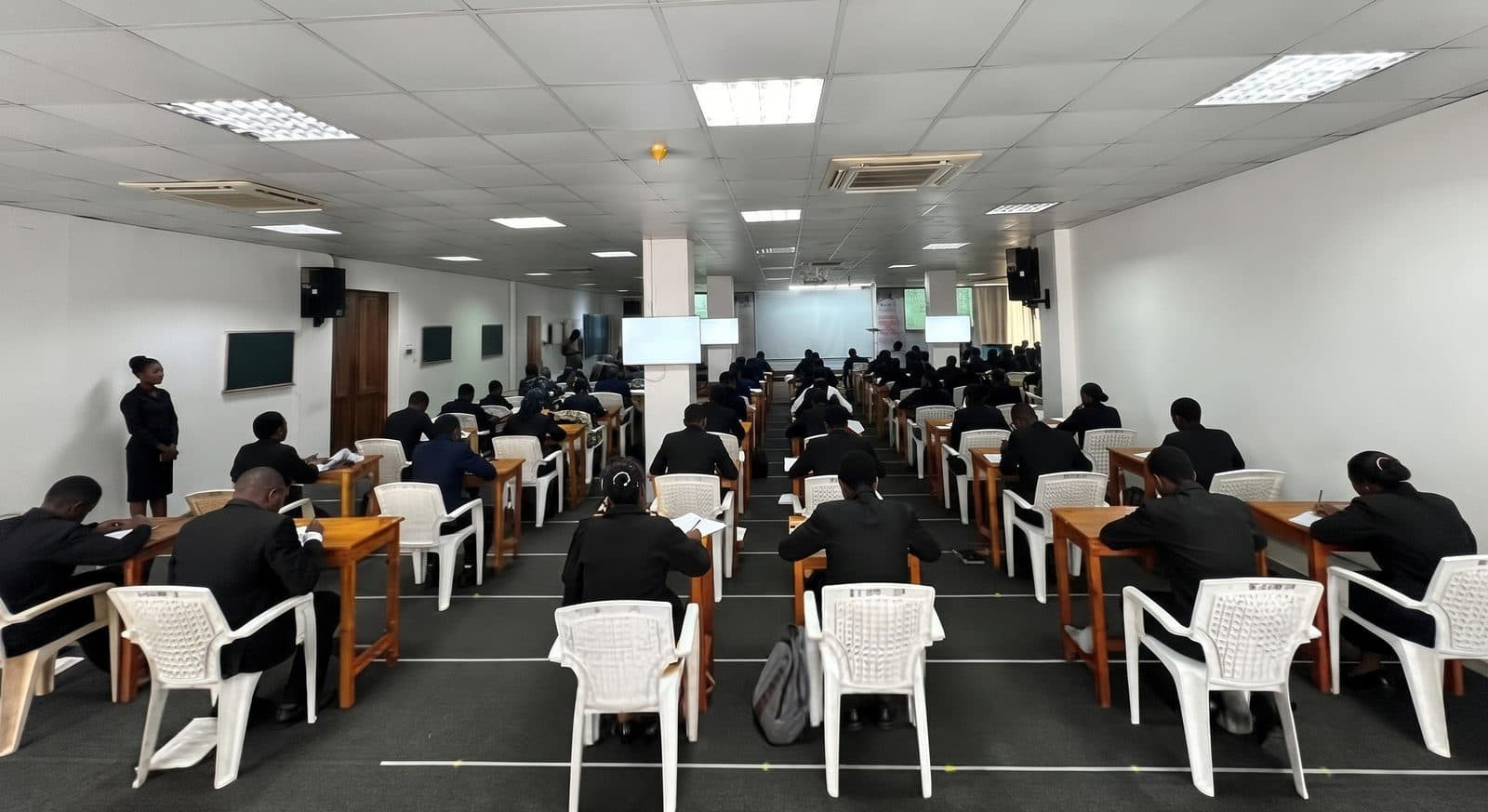Students in dark uniforms writing an exam at rows of wooden desks while an invigilator watches.
